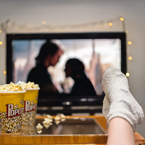 Person watching a movie at home, with a popcorn container on the table and stockinged feet resting on the edge of the table.
