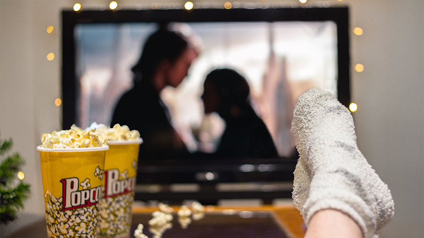 Person watching a movie at home, with a popcorn container on the table and stockinged feet resting on the edge of the table.