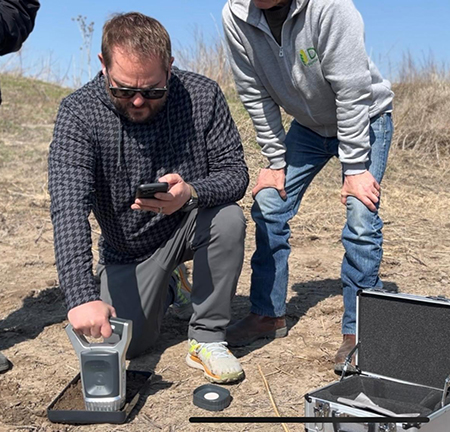 Jonathan Cox, gripping a hand-held device, kneels down to test the soil on a farm in Kyrgyzstan.