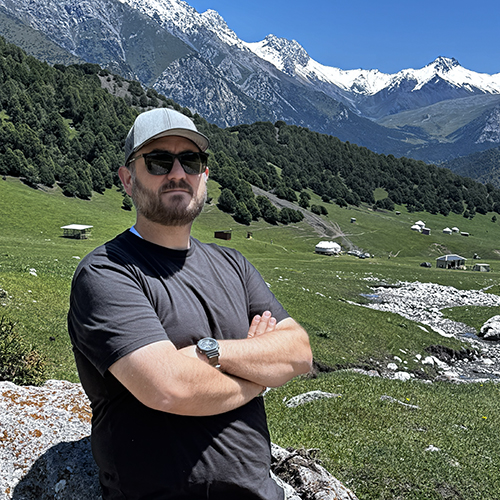 Johnathan Cox with arms folded, with snowy peaked mountains of Kyrgyzstan behind him.