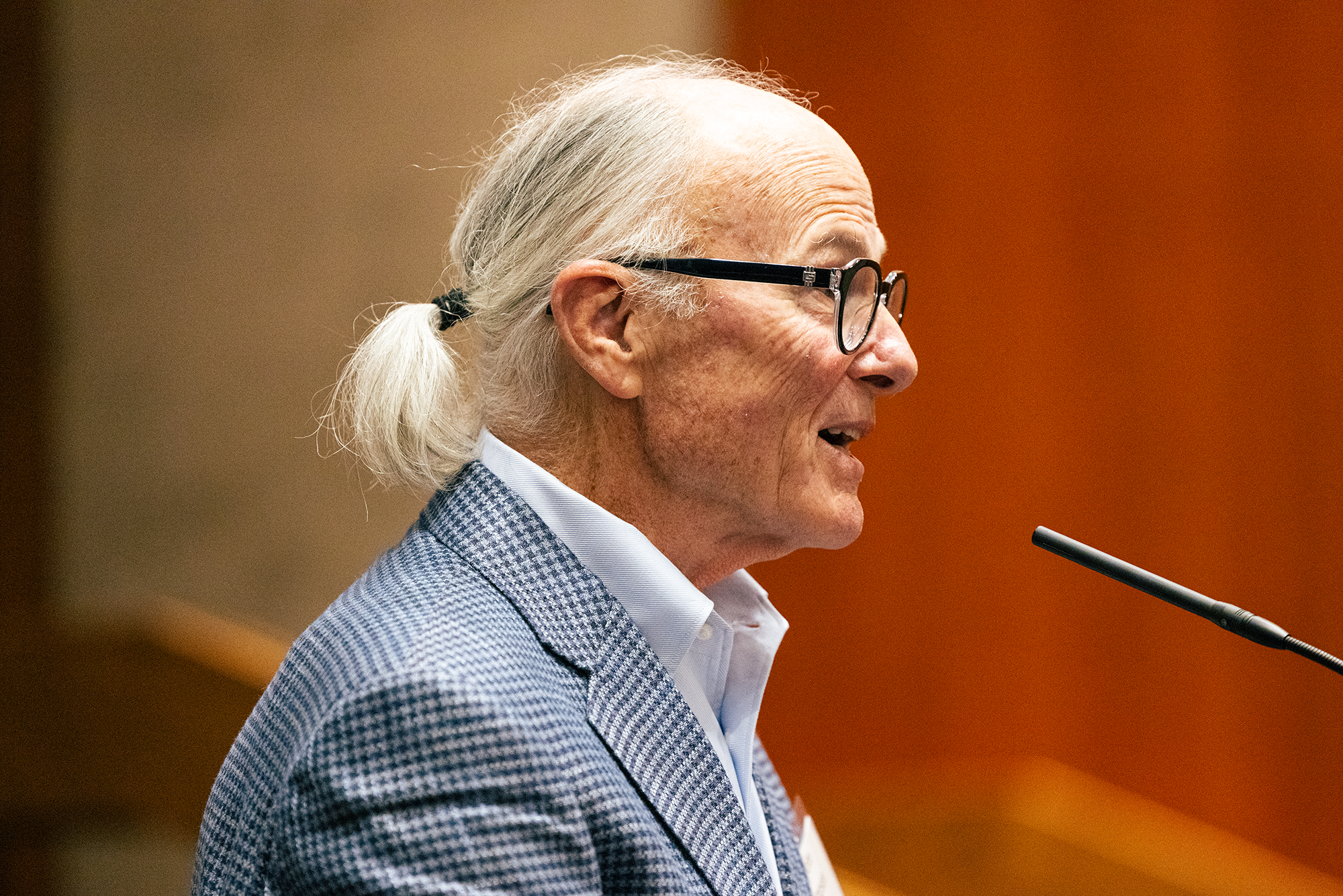 A man with a white ponytail speaks into a microphone on stage