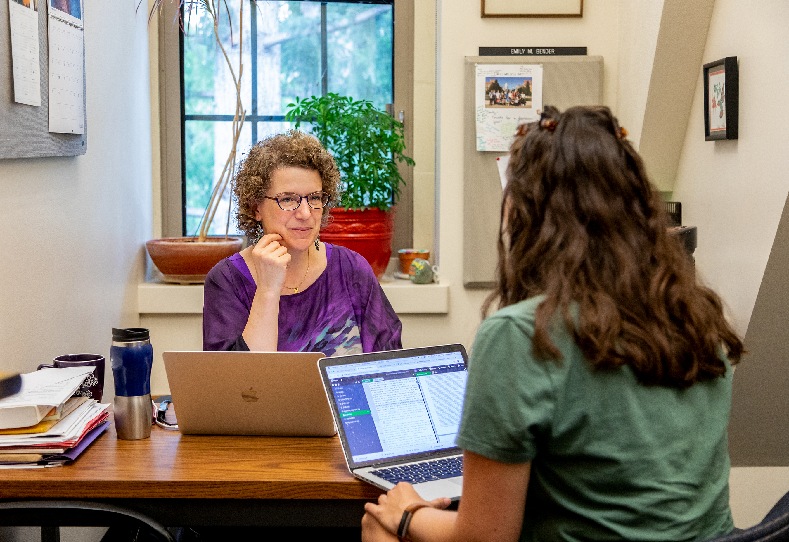 A professor sits behind her desk, chatting with a student. Both of them are holding computers and smiling.
