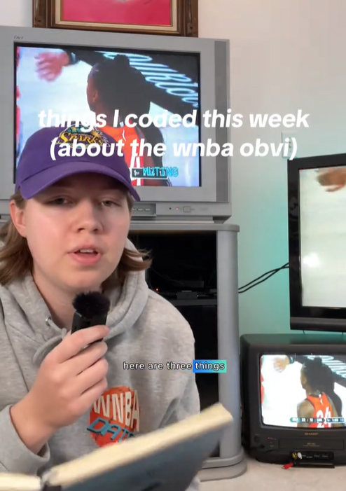 Maddy Brown sitting in front of a TV screen, talking into a handheld mic.