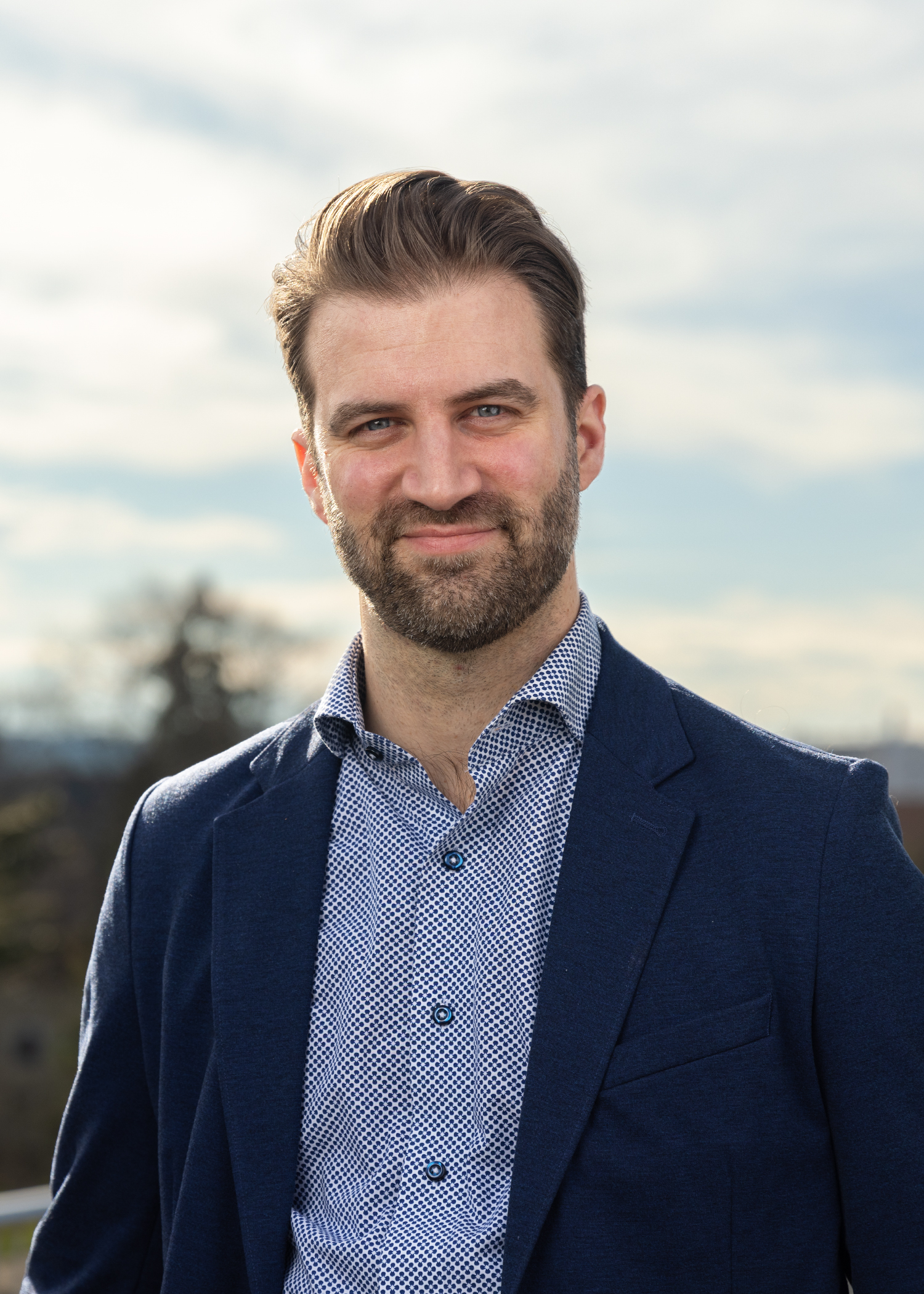 A man wearing a paisley button-down and a blue blazer poses before a cloudy sky.