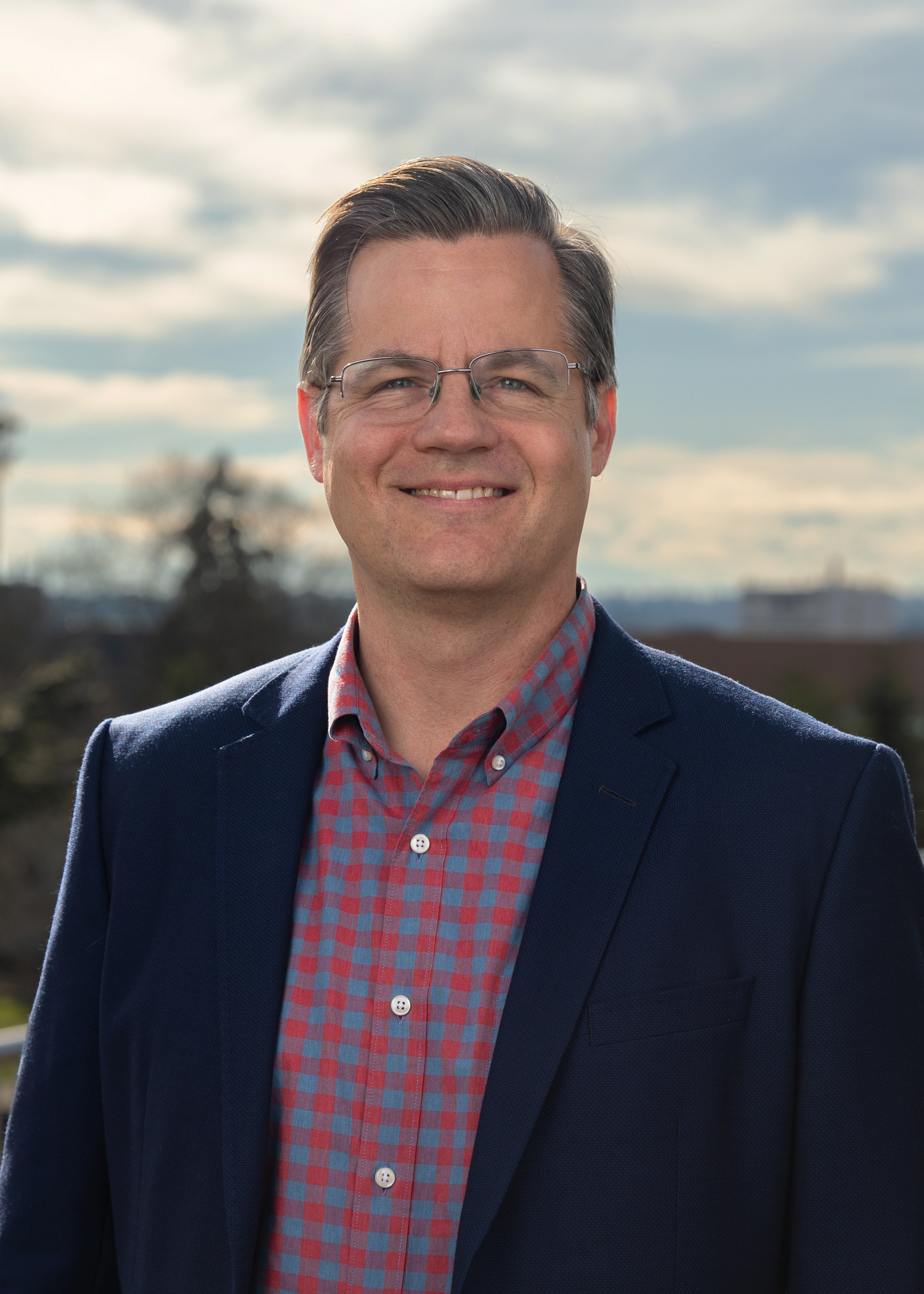 A smiling man in a red flannel button down and blue blazer jacket.