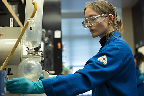 A student wearing a blue lab coat and protective goggles works in a chemistry lab.