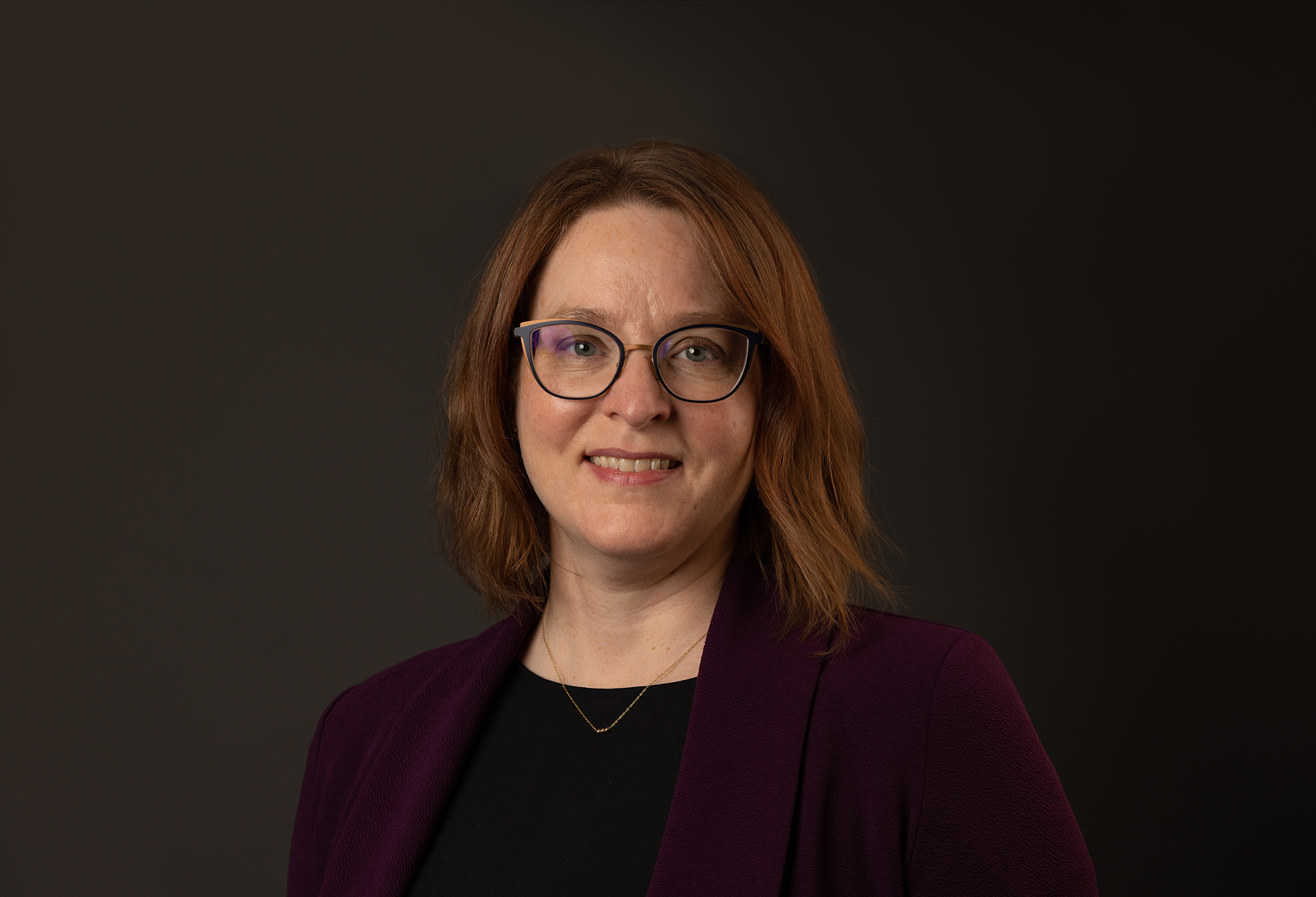 A smiling woman wearing glasses and a maroon blazer.