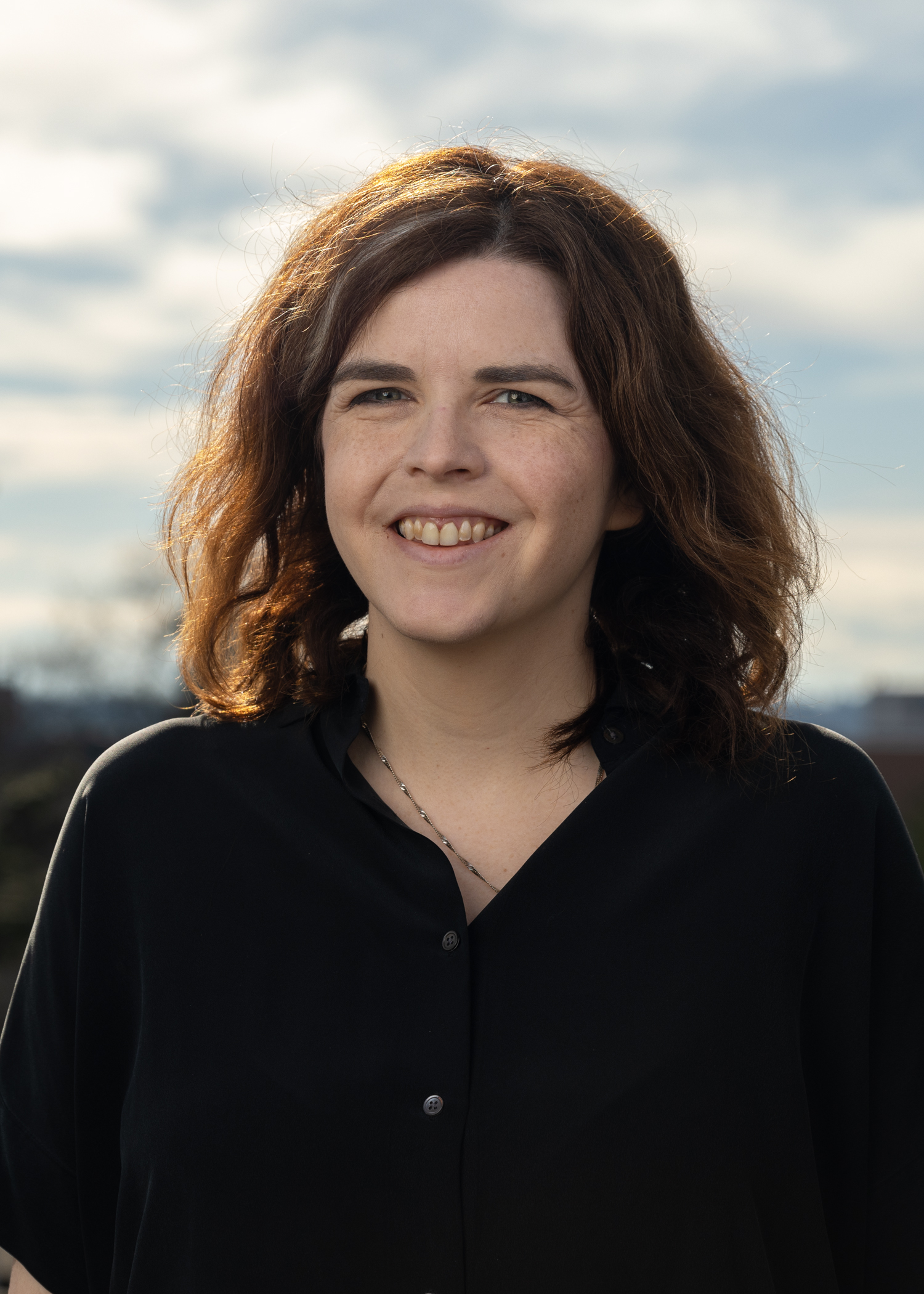 A smiling woman wearing a black button down and delicate chain necklace.