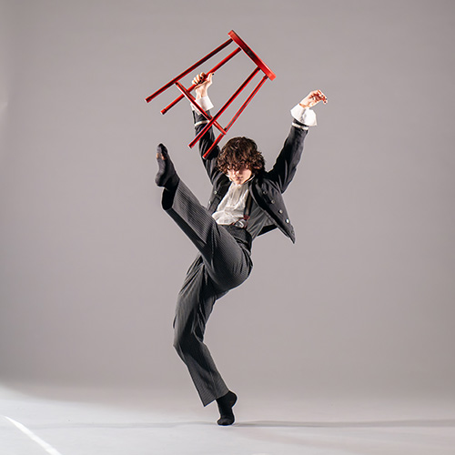 Eddie McClary does a high kick while lifting a red stool overhead during a dance performance.
