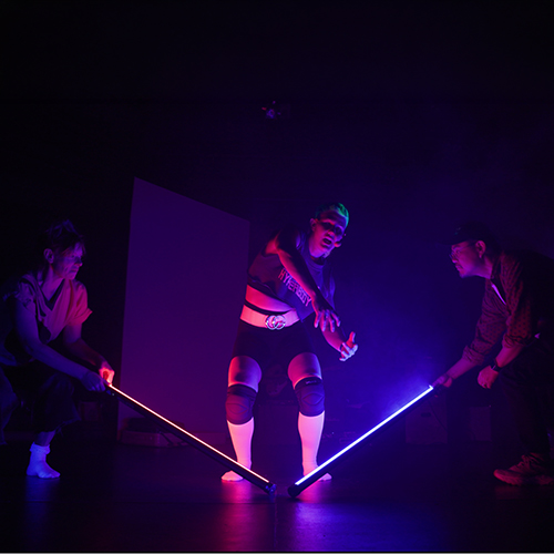 A dancer bathed in purple light dances to music, flanked by two performers who hold light sabers that illuminate the scene.