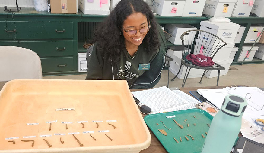 Raquel Matthews sitting behind a long table with small excavated items laid out in even rows. y lab at the Montpelier Museum.