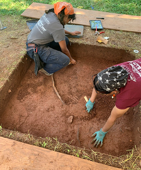 Raquel Matthews and another person excavate inside a small square (about 5 ft. x 5 ft.) dug into the ground. 