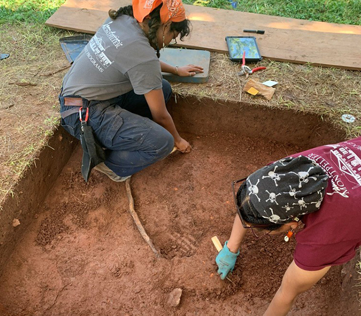 Raquel Matthews and another person excavate inside a small square (about 5 ft. x 5 ft.) dug into the ground. 