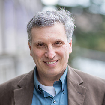 Portrait of Anis Bawarshi wearing a brown jacket and blue shirt in front of a blurry building background