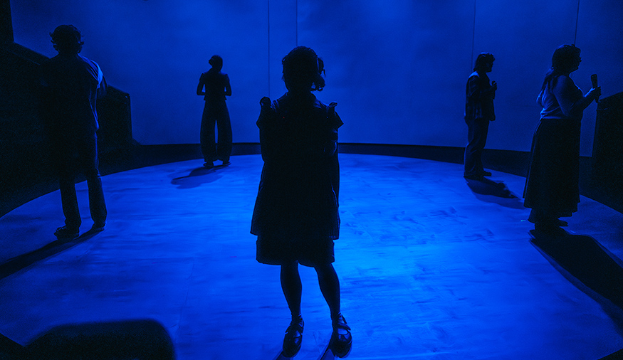 Sihouettes of actors on stage, lit from behind with a blue light. 