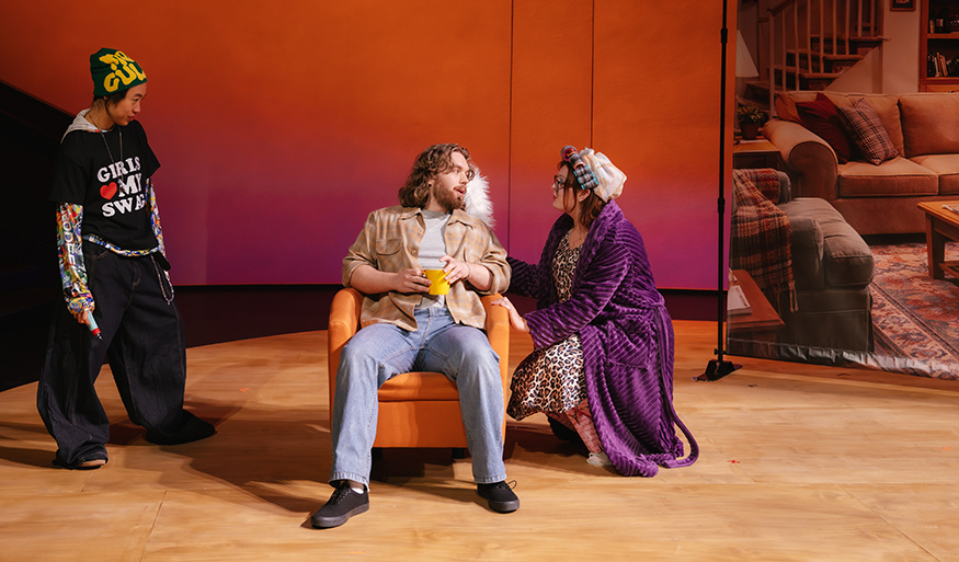 Three actors in contemporary clothes are on stage, with one seated in an upholstered chair. 