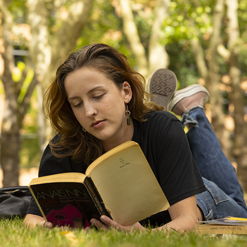 Piper Sorensen laying in the grass, reading an old paperback missing its cover.