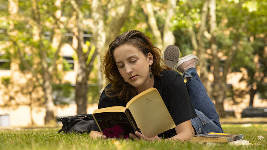 Piper Sorensen laying in the grass, reading an old paperback missing its cover.