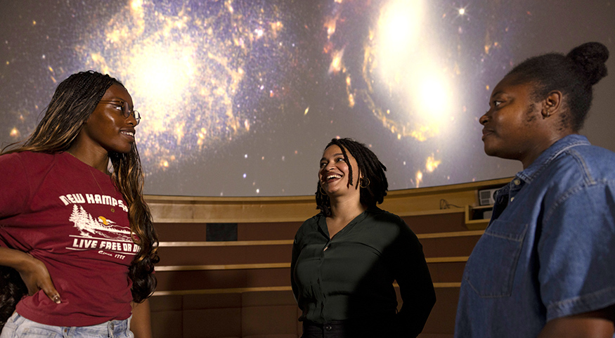 Arianna Long talking with two astronomy students in the UW Planetarium, with galaxies projected on the screen overhead.