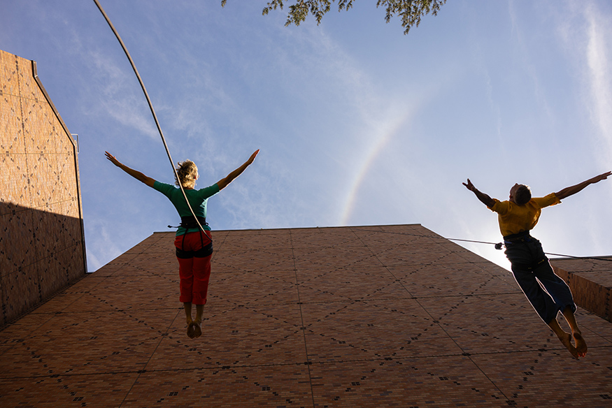 Two dancers with arms spread wide as they dance along a Meany Hall exterior wall, tethered by ropes. 