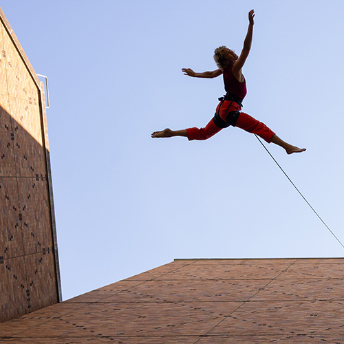 A dancer, roped for safety, leaps skyward off the side of Meany Hall