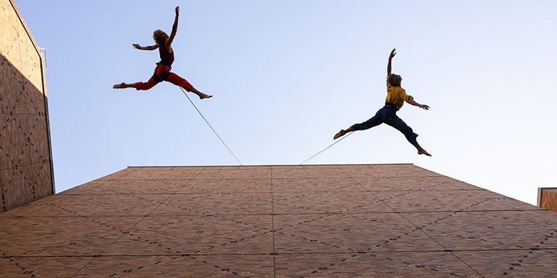 Two dancers in midair as they leap along the side of a tall building.