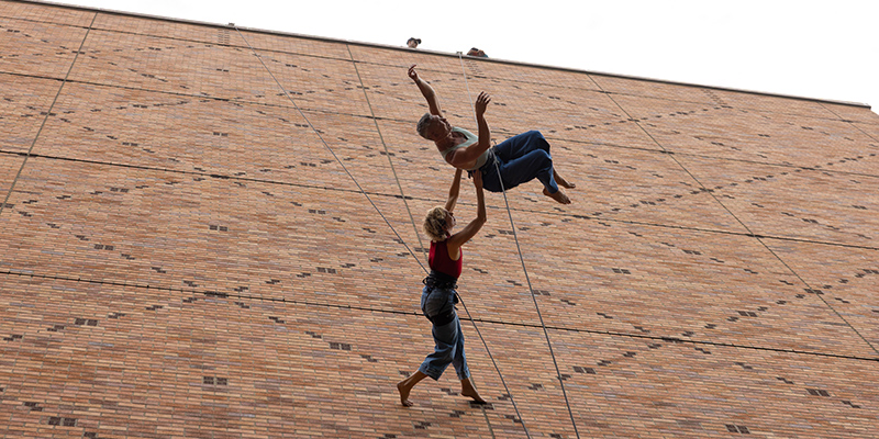 One dancer lifting the other overhead as they dance on the wall of Meany Hall. 