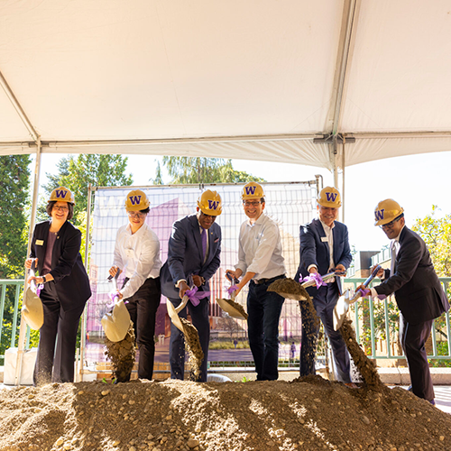 A group of individuals wearing hard hats shovel dirt at a groundbreaking ceremony.