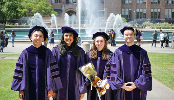 Four students in cap and gown in front of Drumheller Fountain.